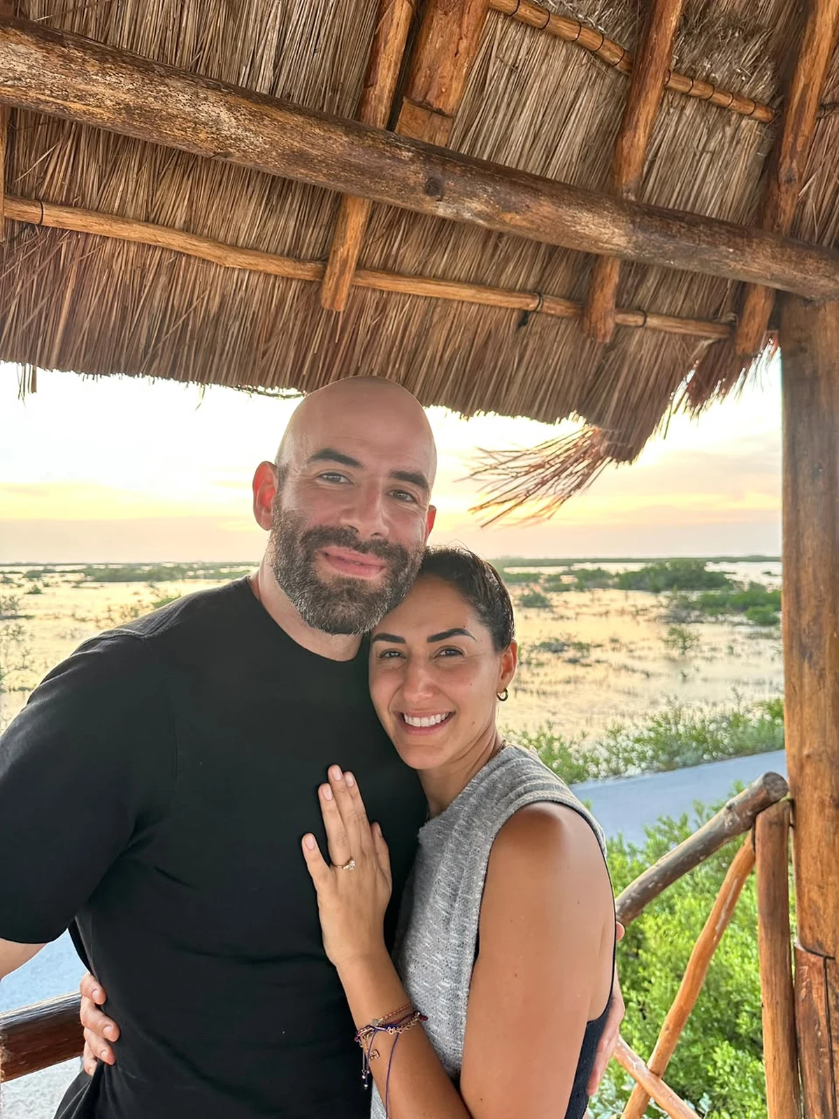 Nora and Marc together under a palapa at sunset, with the water and mangroves behind them