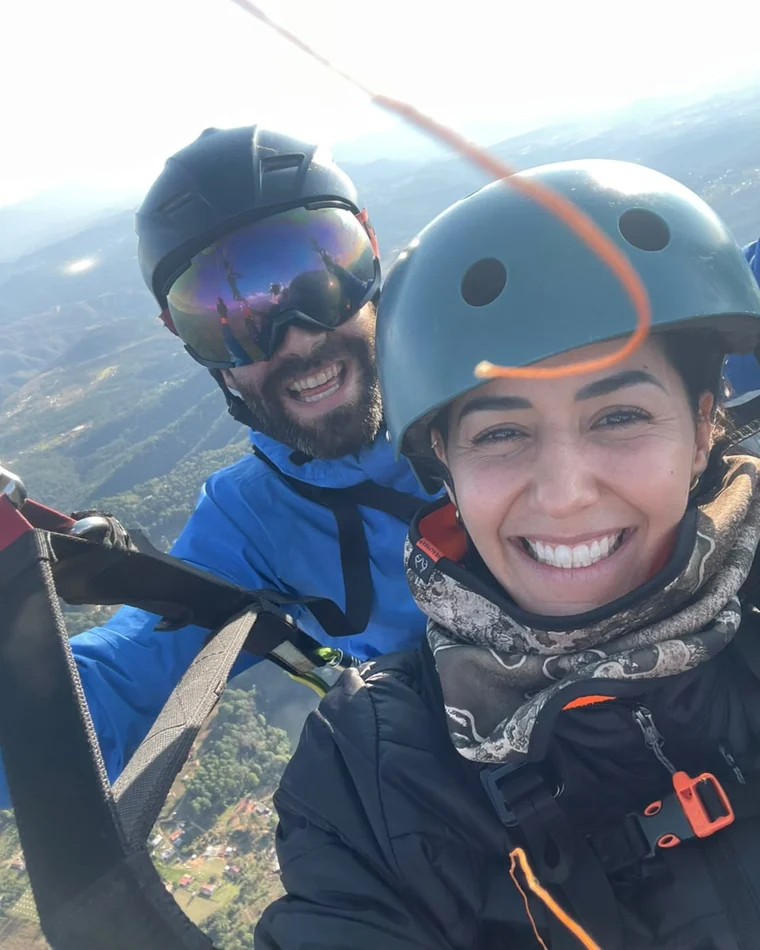 Nora and Marc paragliding in helmets, grinning, with green hills far below